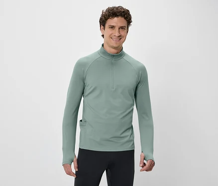 Un homme aux cheveux bouclés porte un t-shirt vert à manches longues avec fermeture éclair et un pantalon noir sur fond blanc.