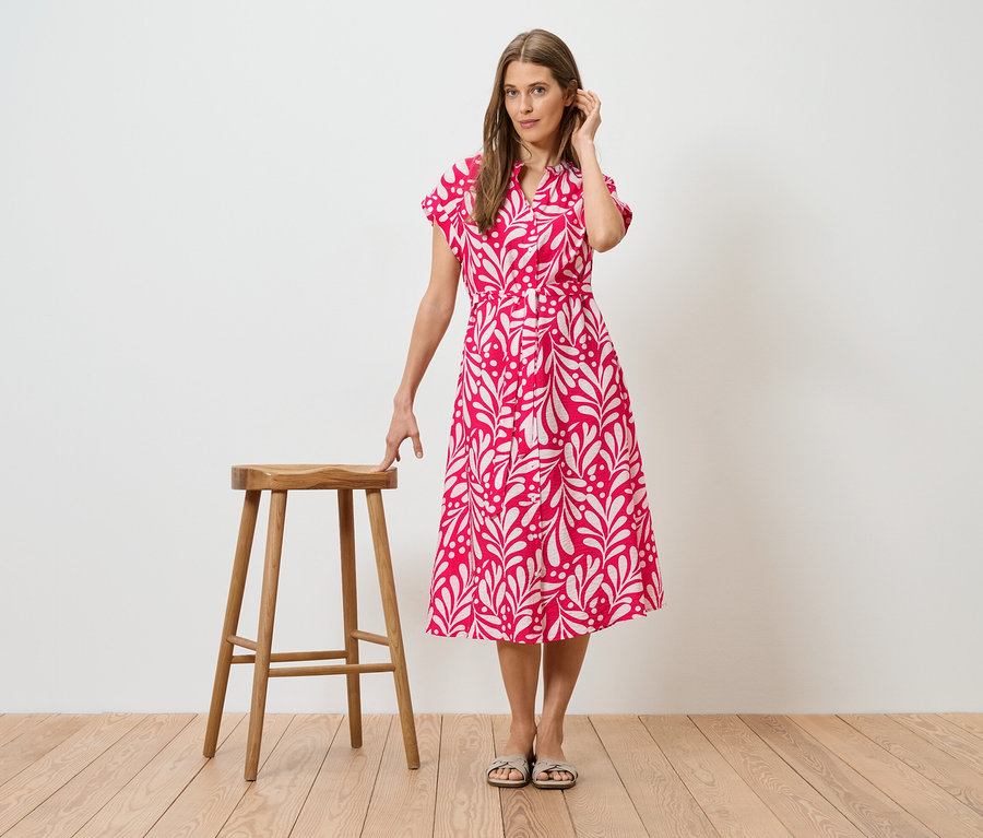 Femme en robe rose à motif de feuilles blanches, debout à côté d'un tabouret en bois.