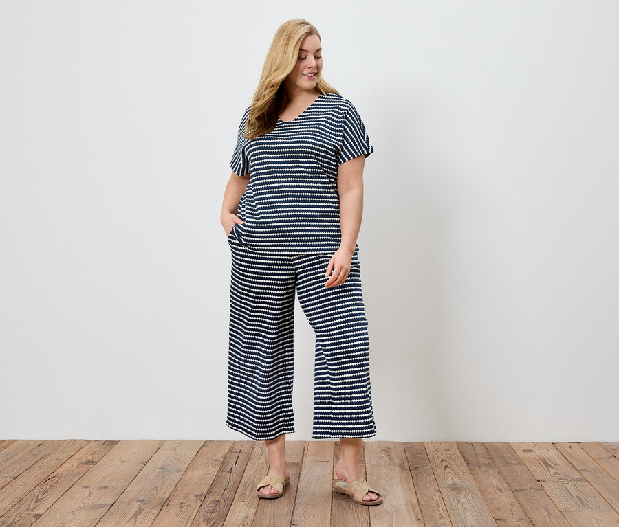 Une femme aux cheveux blonds porte un pyjama rayé bleu marine et blanc et des sandales devant un mur blanc.