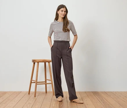 Une femme portant un haut rayé et un pantalon marron se tient à côté d'un tabouret sur un plancher en bois.