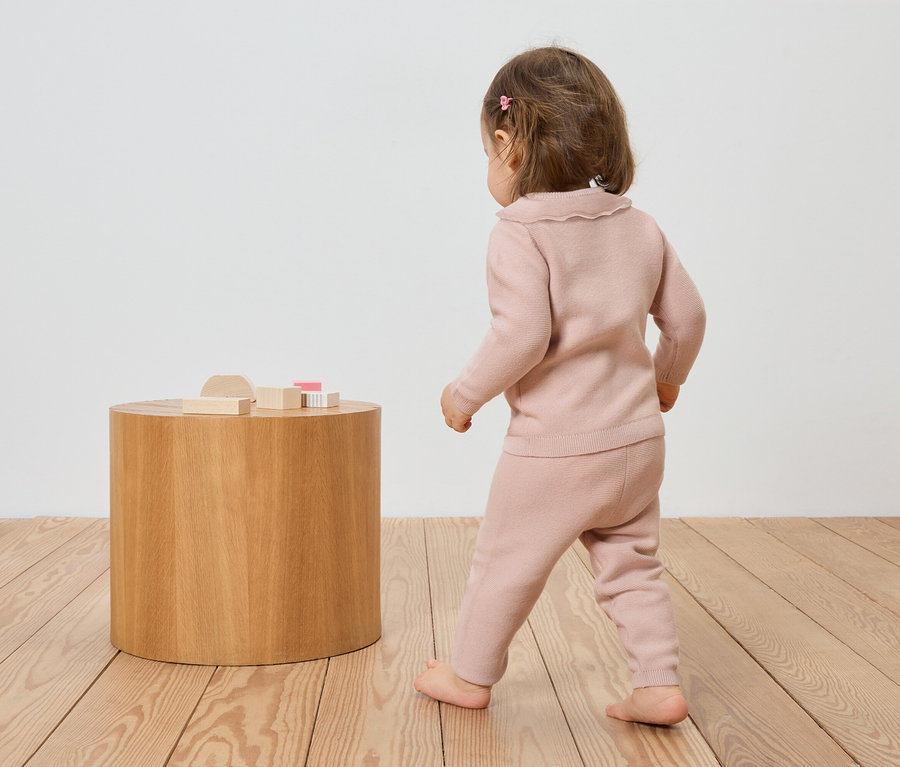 Un jeune enfant en tricot rose marche pieds nus sur un plancher en bois vers une table ronde en bois avec des jouets en bois.