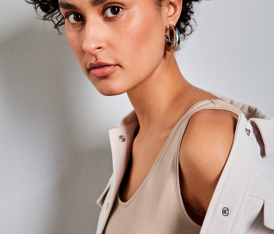 Femme aux cheveux courts et bouclés portant un haut classique Street One beige et une veste claire sur les épaules, regardant l'appareil photo.