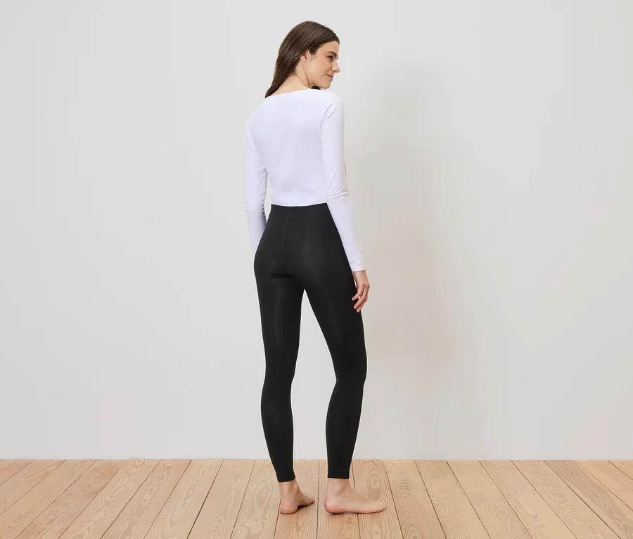 Une femme portant un haut blanc et un legging noir se tient sur un plancher en bois devant un mur blanc et regarde par-dessus son épaule.