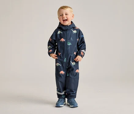 Un garçon souriant se tient sur un fond clair, vêtu d'une combinaison bleu marine à motifs de dinosaures et de chaussures bleues.