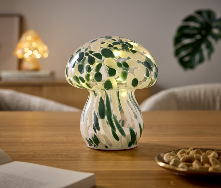 Une lampe en forme de champignon avec un motif vert et blanc et des lumières est posée sur une table en bois. À côté, il y a un livre ouvert et une petite assiette de noix de cajou. À l'arrière-plan, une autre lampe champignon et une plante sont visibles.
