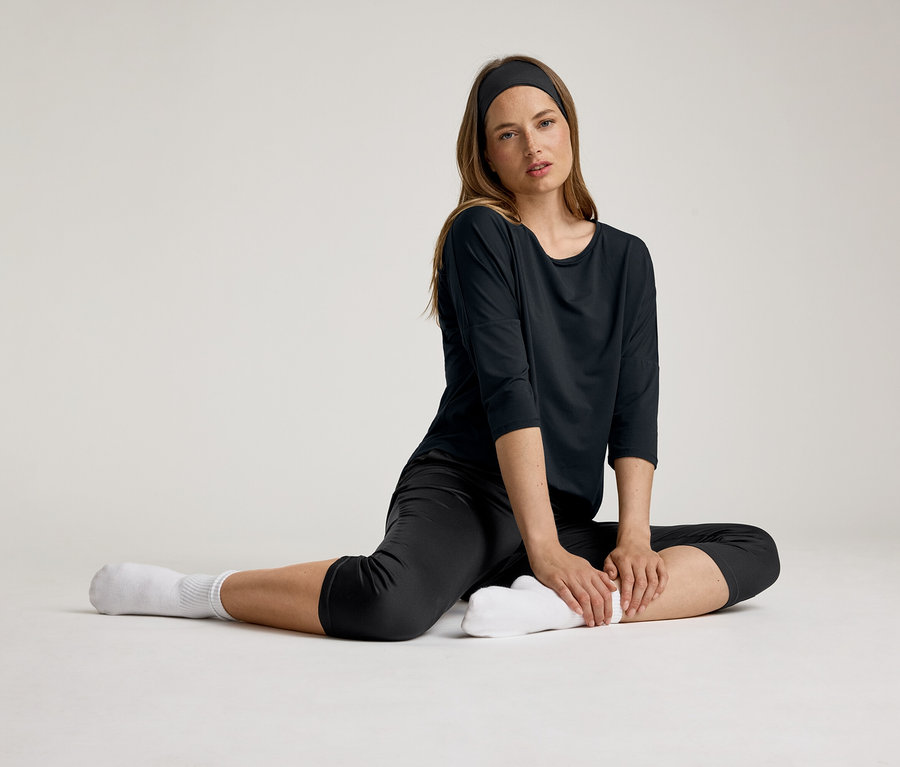 Une femme est assise et porte un t-shirt de sport 3/4 noir. Elle porte un bandeau et des chaussettes blanches.