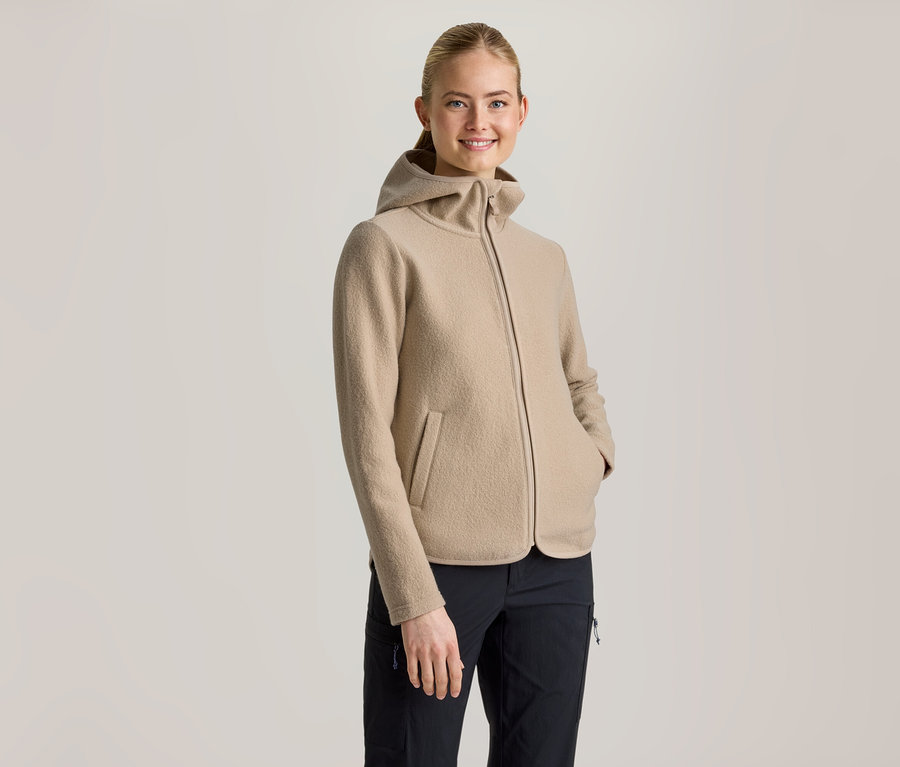 Une femme souriante aux cheveux attachés porte une veste à capuche beige et un pantalon noir, les mains dans les poches de la veste.