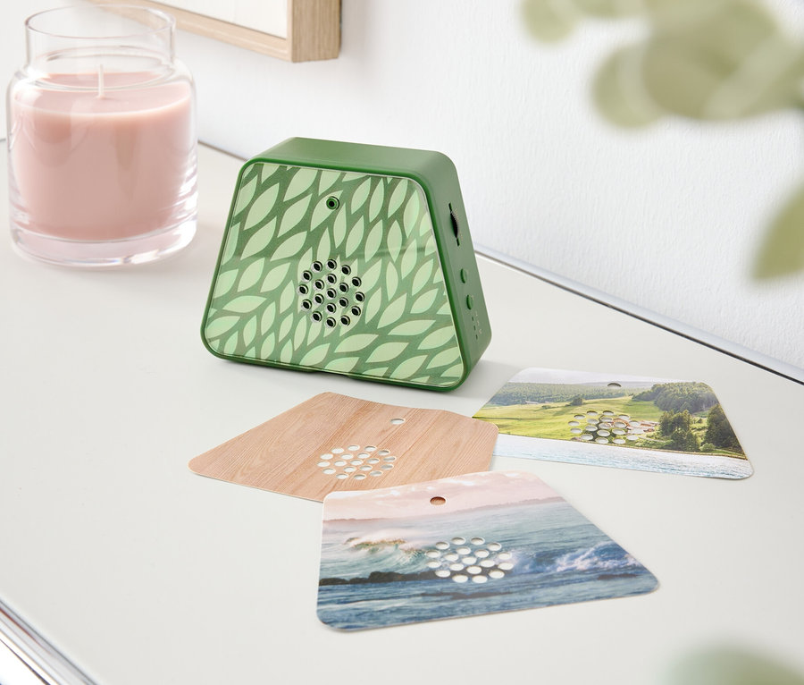 Sur une table, il y a une bougie rose, un diffuseur de parfum vert avec un motif de feuilles et trois cartes de parfum.