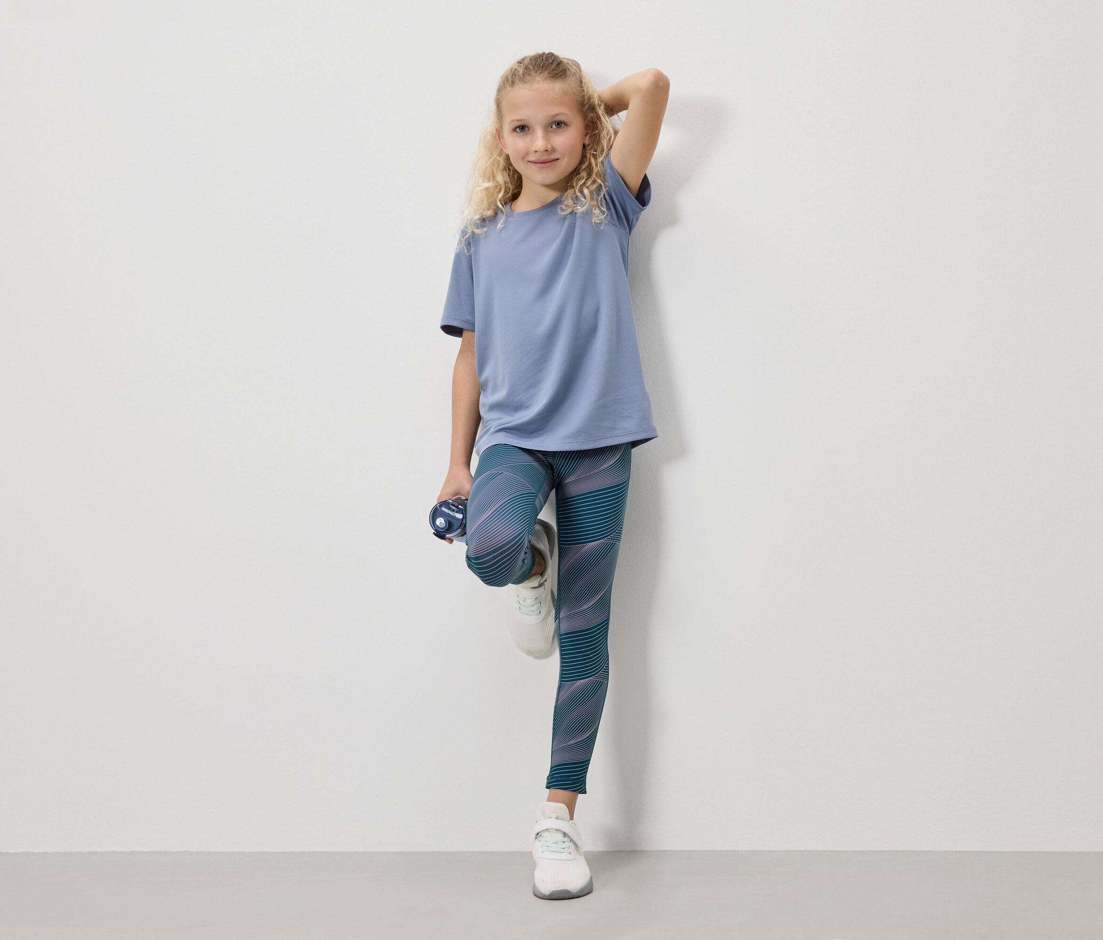 Une fille blonde portant un t-shirt bleu, des leggings et des baskets blanches se tient avec une bouteille d'eau devant un mur blanc.