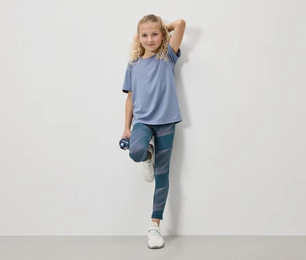 Une fille blonde portant un t-shirt bleu, des leggings et des baskets blanches se tient avec une bouteille d'eau devant un mur blanc.