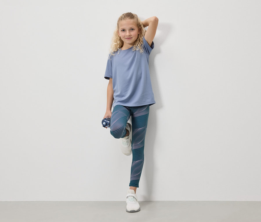 Une fille blonde portant un t-shirt bleu, des leggings et des baskets blanches se tient avec une bouteille d'eau devant un mur blanc.