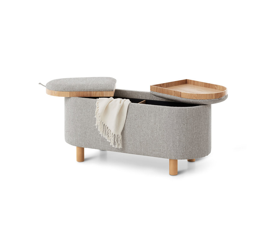 Banc de rangement rembourré gris clair avec pieds en bois et deux couvercles ouverts, l'un rembourré et l'autre un plateau en bois. Une couverture crème pend sur le côté.