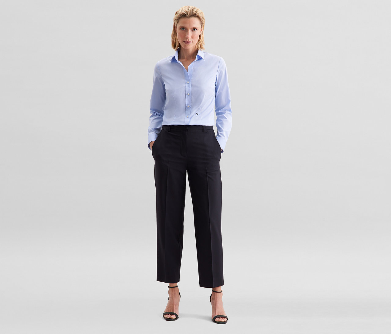 Femme debout, les mains dans les poches, portant un chemisier Slim Fit non iron Seidensticker bleu clair, un pantalon noir et des sandales noires à talons.