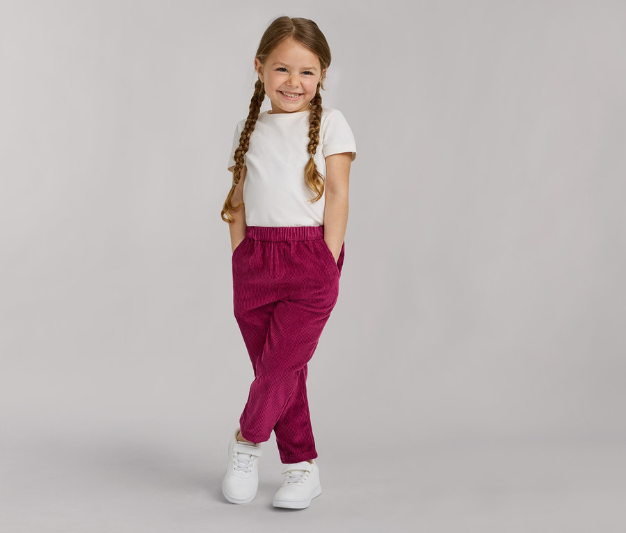 Une jeune fille souriante avec des tresses, portant un t-shirt blanc, un pantalon en velours côtelé magenta et des baskets blanches, les mains dans les poches.