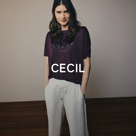 Une femme aux cheveux foncés porte un t-shirt violet avec une inscription et un pantalon clair à cordon de serrage. Elle se tient les mains dans les poches et regarde l'appareil photo.