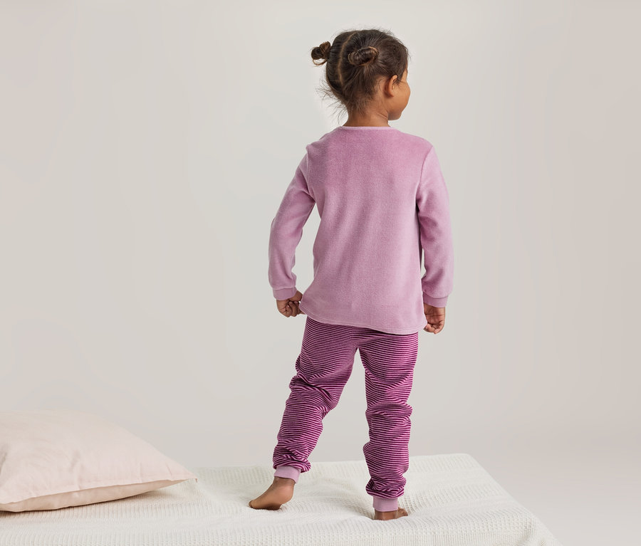 Enfant vu de dos, en pyjama rose avec un pantalon rayé, debout sur une couverture blanche à côté d'un oreiller rose clair.