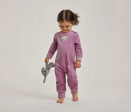 Un tout-petit souriant, vêtu d'une grenouillère en velours violette avec un visage de chat sur la poitrine, tient un animal en peluche gris et regarde vers le bas, pieds nus.