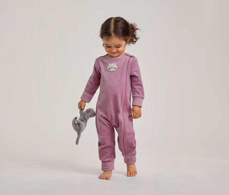 Un tout-petit souriant, vêtu d'une grenouillère en velours violette avec un visage de chat sur la poitrine, tient un animal en peluche gris et regarde vers le bas, pieds nus.