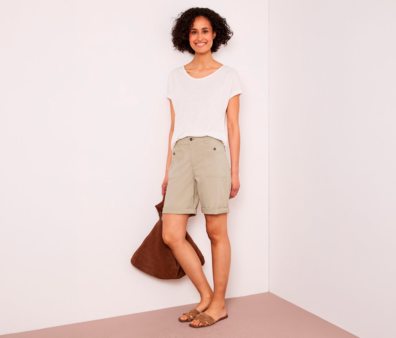 Femme souriante aux cheveux courts et bouclés portant un t-shirt blanc, un bermuda Street One « Yulius » beige et des sandales marron, tenant un sac marron.