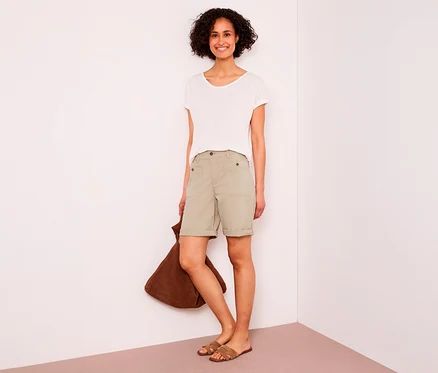 Femme souriante aux cheveux courts et bouclés portant un t-shirt blanc, un bermuda Street One « Yulius » beige et des sandales marron, tenant un sac marron.