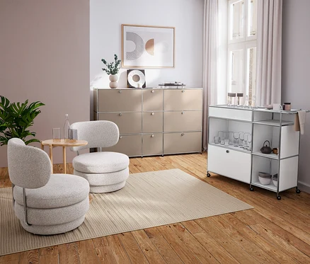 Salon moderne avec deux fauteuils gris clair, une table d'appoint ronde en bois, un buffet beige et un chariot de service blanc.