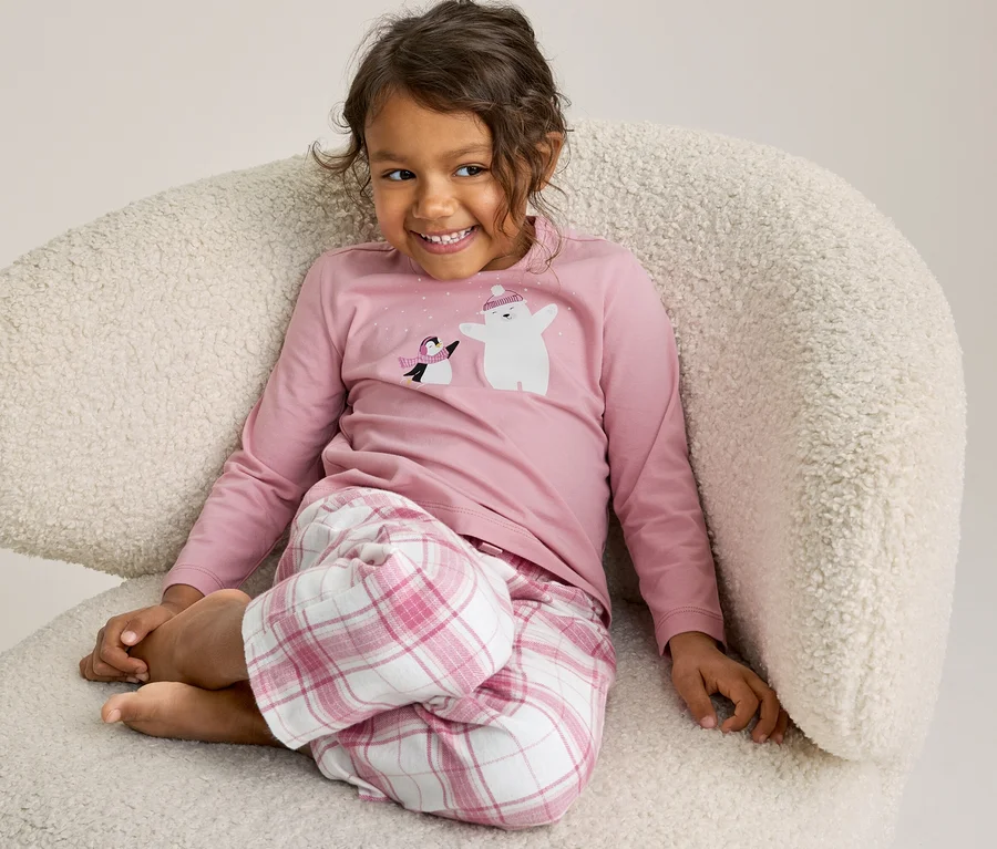 Jeune fille aux cheveux bouclés assise sur un fauteuil blanc moelleux, portant un pyjama rose avec un ours polaire et un pingouin, souriante.