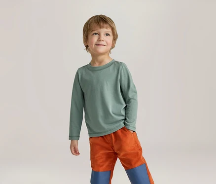 Un jeune garçon aux cheveux châtains clairs porte un t-shirt vert à manches longues et un pantalon orange avec des empiècements bleus. Il sourit en regardant vers le haut.