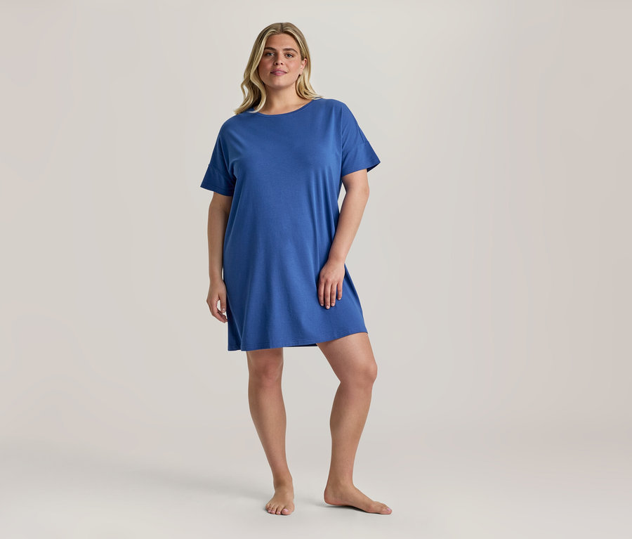 Une femme blonde est debout pieds nus, portant un Nachthemd bleu à manches courtes qui arrive au-dessus des genoux.