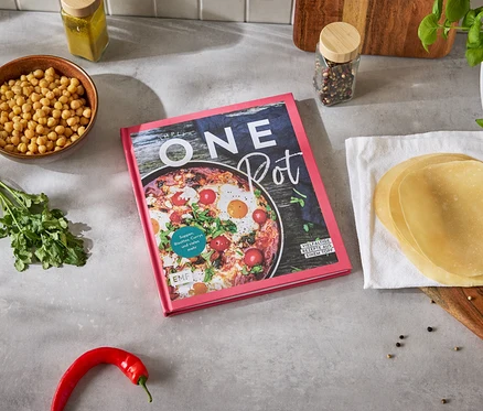 Un livre de cuisine 'One Pot' est posé sur un plan de travail de cuisine, entouré de pois chiches, de coriandre, de piment et de tortillas.