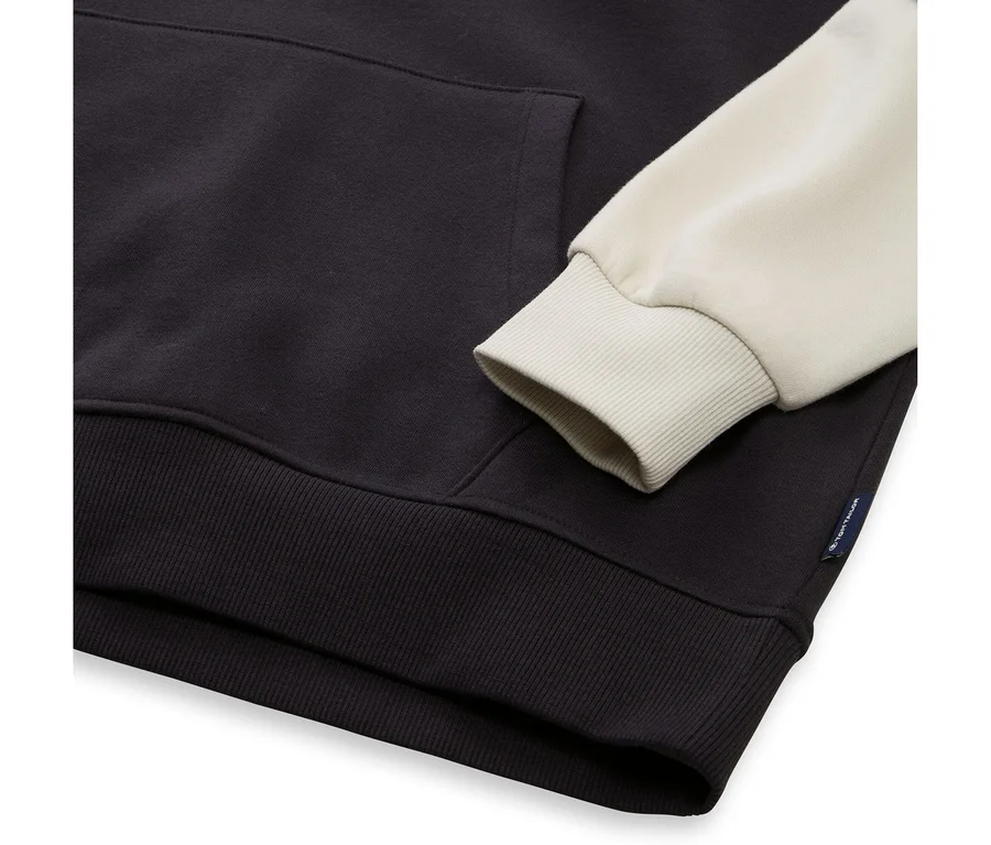 Gros plan d'un sweat à capuche bicolore avec un corps bleu foncé et une manche crème clair. Il présente une poche kangourou, des poignets et un ourlet côtelés. Une petite étiquette noire avec du texte blanc est visible sur le côté.