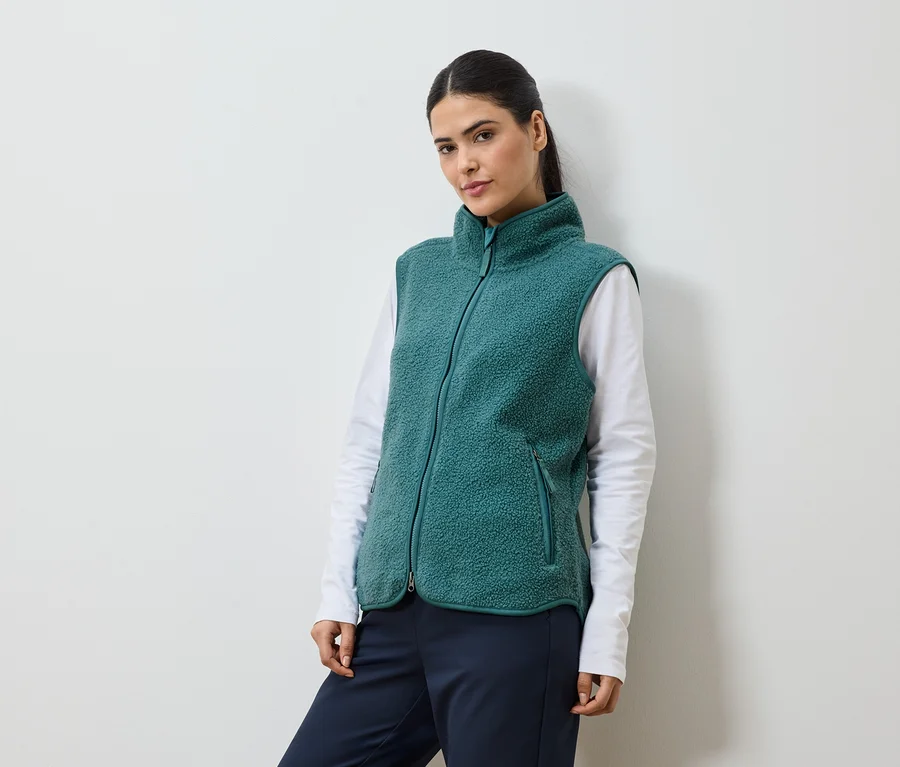 Femme avec queue de cheval portant un gilet en polaire vert sur un t-shirt blanc à manches longues et un pantalon bleu marine.