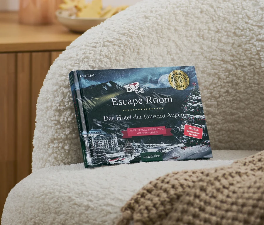 Sur un fauteuil se trouvent un livre calendrier de l’Avent Escape Room « Das Hotel der tausend Augen » (L’hôtel aux milles yeux, en allemand) et une couverture beige.