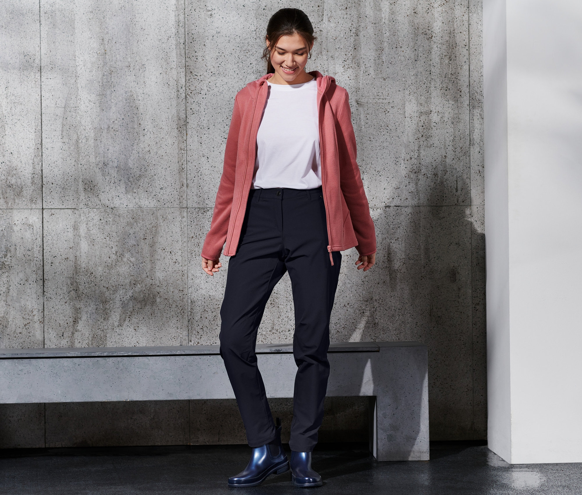 Une femme souriante porte un pantalon softshell, un t-shirt blanc et un sweat à capuche rose.