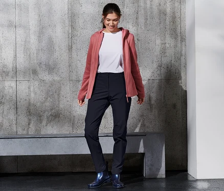 Une femme souriante porte un pantalon softshell, un t-shirt blanc et un sweat à capuche rose.