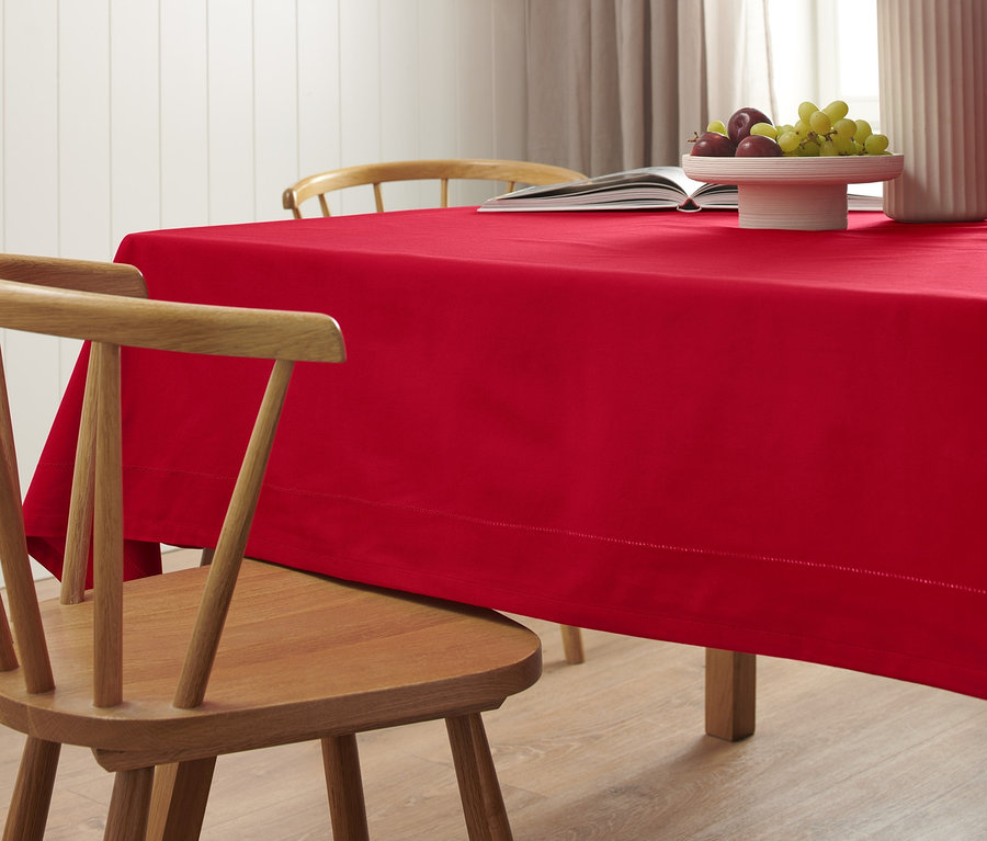 Une nappe rouge de grande taille est posée sur une table en bois. Un livre ouvert, un vase et un saladier de fruits sont posés sur la nappe.
