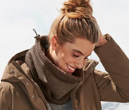 Une femme souriante portant un chignon, une écharpe-tube en maille polaire marron et une veste marron. Des montagnes sont visibles en arrière-plan.