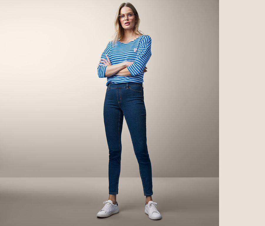 Une femme blonde pose les bras croisés, portant un jegging, un haut rayé et des baskets blanches.