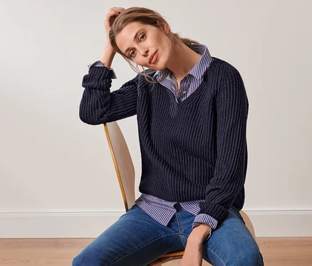 Une femme est assise sur une chaise et porte une chemise rayée et un pull en grosse maille bleu foncé avec de la laine.