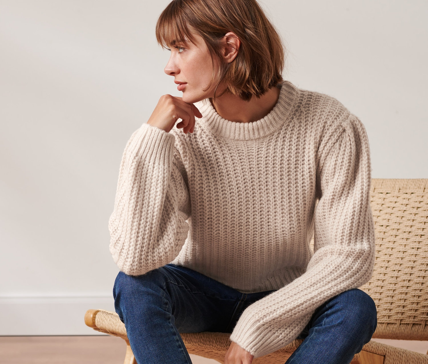 Une femme est assise sur une chaise et porte un pull en grosse maille crème avec de la laine, un jean bleu et des bottes noires.
