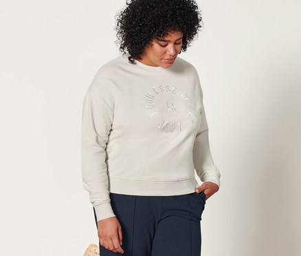Une femme se tient à côté d'une chaise en osier et porte un sweat-shirt crème et un pantalon de jogging bleu foncé à nervures.