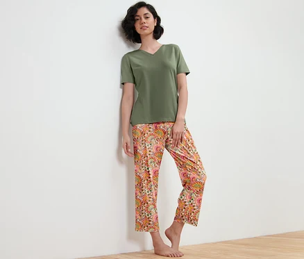 Une femme en pyjama vert avec un pantalon à fleurs est appuyée contre un mur blanc.