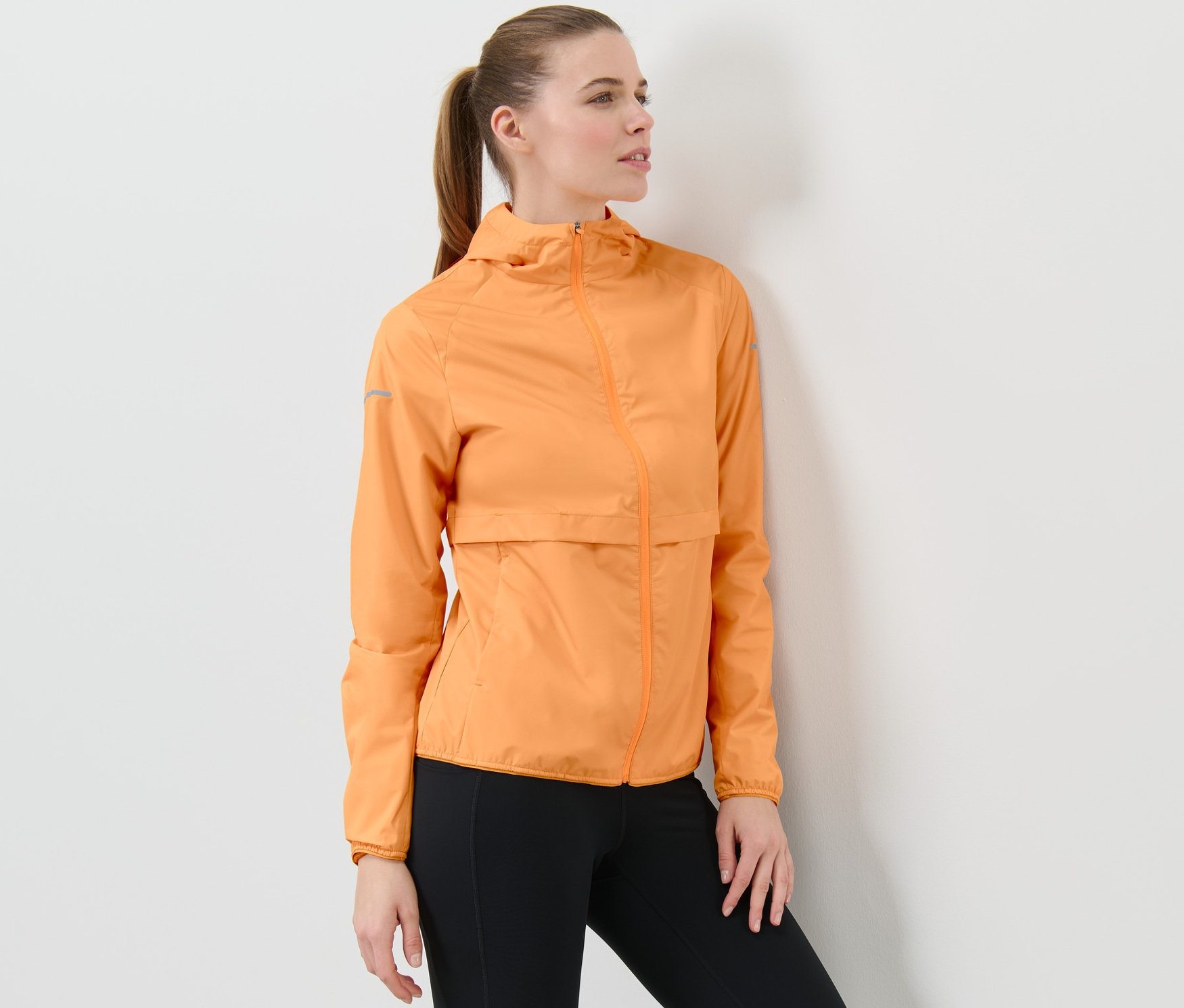 Femme posant et portant une veste de course coupe-vent orange fluo et un legging noir.