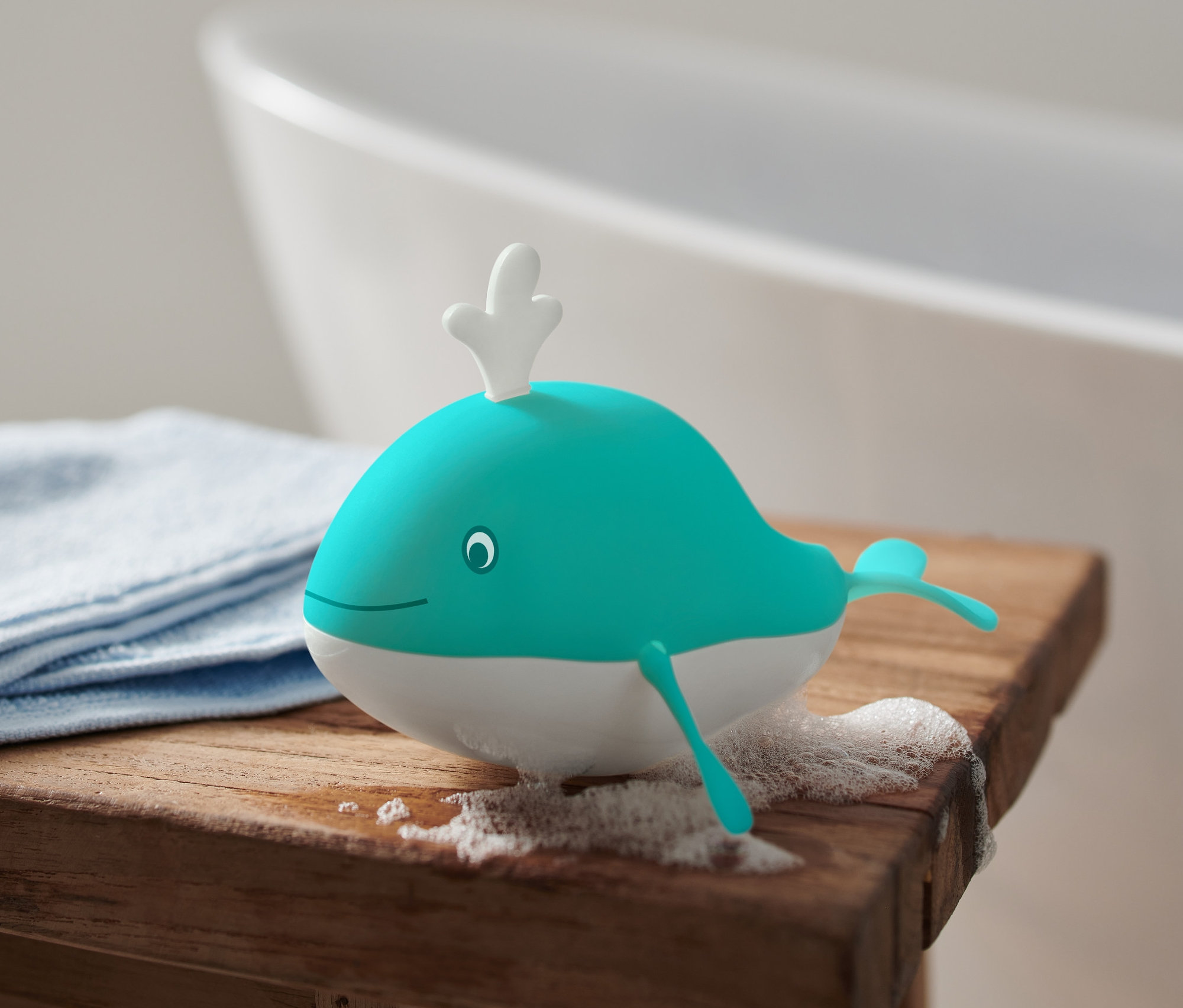 Un jouet de bain Baleine à remonter se trouve sur un tabouret en bois à côté d'un tissu bleu et de la mousse, une baignoire en arrière-plan.