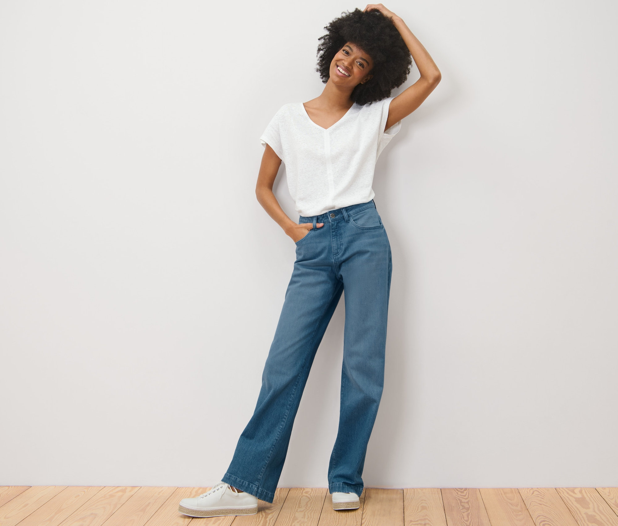 Une femme avec une coupe afro pose en jean palazzo « Neni » et un t-shirt blanc contre un mur blanc.