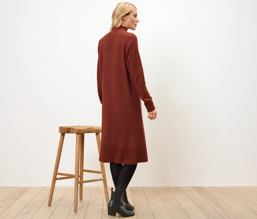 Une femme se tient de côté et porte une robe en maille avec teneur en laine, des collants noirs et des bottines. Un tabouret en bois se trouve à côté d'elle.