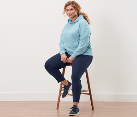 Une femme est assise sur un tabouret et porte un sweat-shirt à capuche bleu clair, un pantalon stretch bleu foncé et des tennis slip-on.