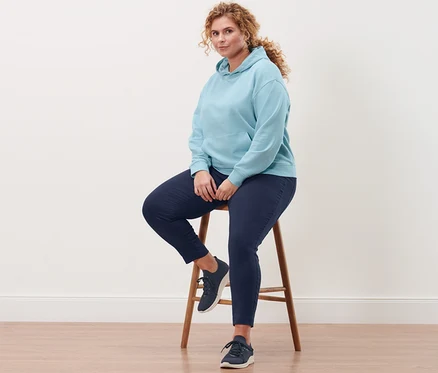 Une femme est assise sur un tabouret et porte un sweat-shirt à capuche bleu clair, un pantalon stretch bleu foncé et des tennis slip-on.