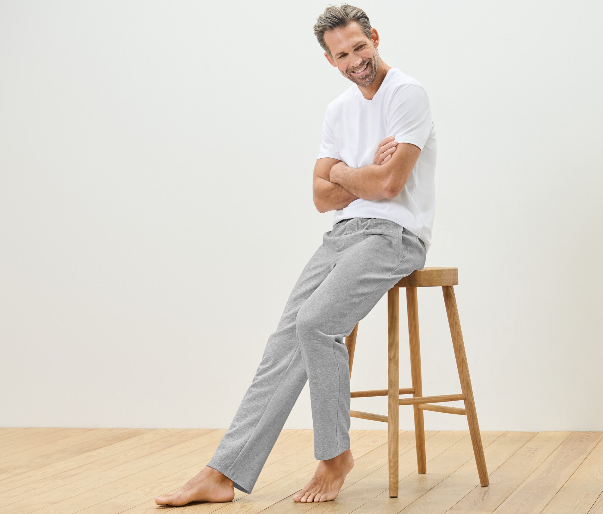 Un homme est assis sur un tabouret en bois et porte un pantalon d’intérieur et un t-shirt.