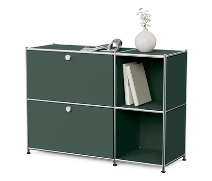 Un buffet en métal « CN3 » vert olive avec 2 abattants, sur lequel repose un journal. À côté se trouve une étagère ouverte avec des livres et un vase.