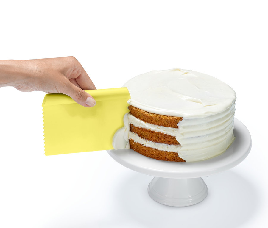 Une main utilise un grattoir jaune du lot d'ustensiles de pâtisserie pour appliquer un glaçage blanc sur les couches d'un gâteau.